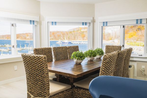 A dining room with a wooden table, woven chairs, and potted plants. Large windows offer a view of a scenic waterfront and autumn foliage ending the sentence.