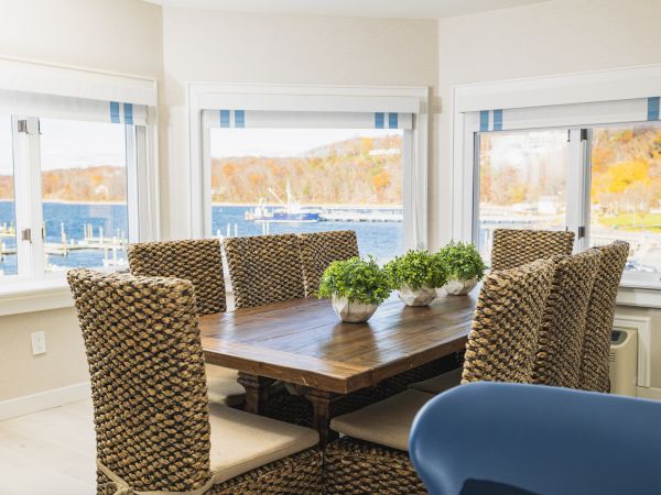 A dining room with a wooden table, woven chairs, and potted plants. Large windows offer a view of a scenic waterfront and autumn foliage ending the sentence.
