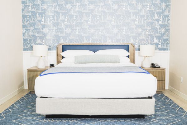 A neatly arranged bedroom with a bed, two bedside tables, and lamps, against a nautical-themed wallpaper and a blue rug on the floor.