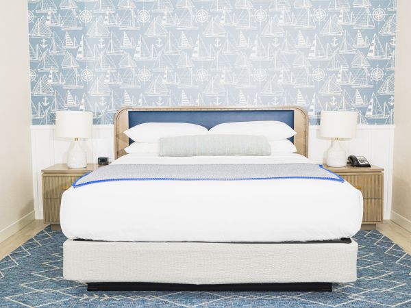 A neatly arranged bedroom with a bed, two bedside tables, and lamps, against a nautical-themed wallpaper and a blue rug on the floor.