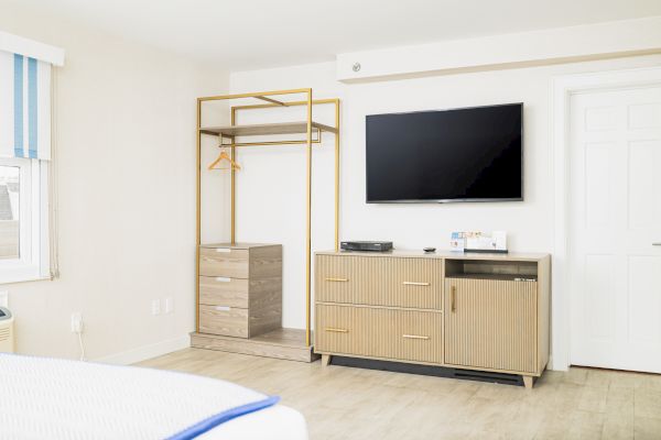 The image shows a modern, minimalistic room with a TV mounted on the wall, a cabinet below, an open closet, wooden floor, and a window with blinds.