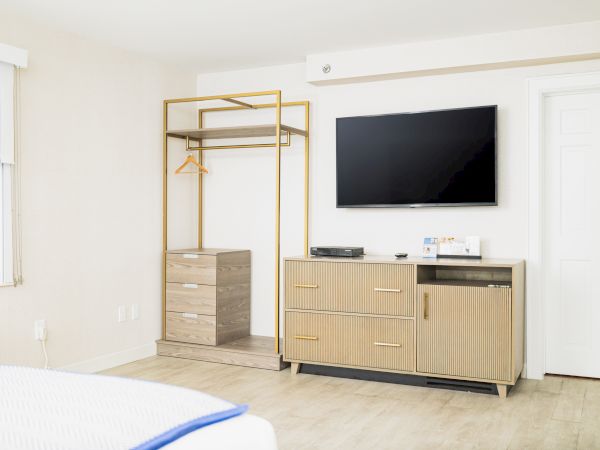 The image shows a modern, minimalistic room with a TV mounted on the wall, a cabinet below, an open closet, wooden floor, and a window with blinds.
