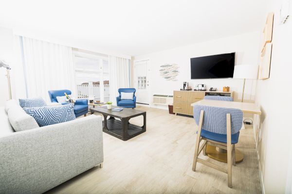 Bright living room with grey sofa, blue chairs, TV, a desk, and a coffee table on wooden floors. Curtains allow natural light into the space.