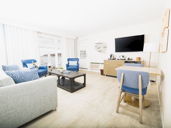 Bright living room with grey sofa, blue chairs, TV, a desk, and a coffee table on wooden floors. Curtains allow natural light into the space.