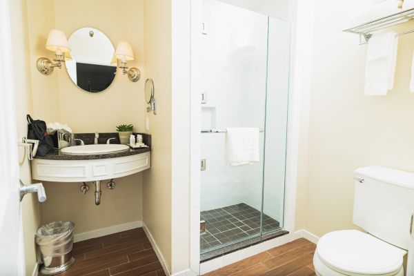 A modern bathroom with a circular mirror, sink, glass shower, toilet, and towel rack. The space is clean and well-lit by wall lamps.