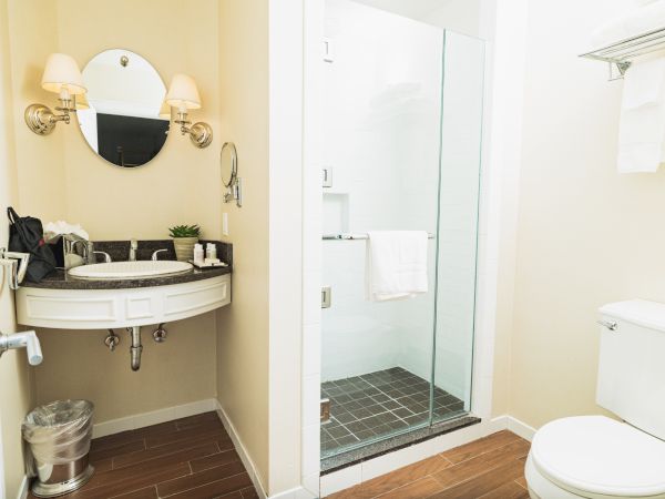 A modern bathroom with a circular mirror, sink, glass shower, toilet, and towel rack. The space is clean and well-lit by wall lamps.