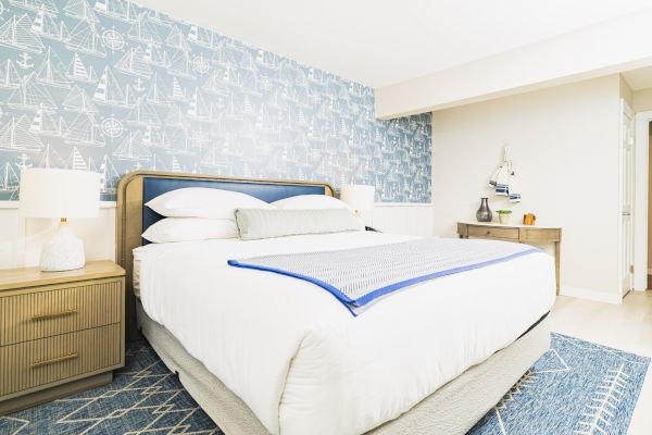 A cozy bedroom with a nautical theme, featuring a bed with white linens, a nightstand with a lamp, and a blue patterned accent wall with sailboats.