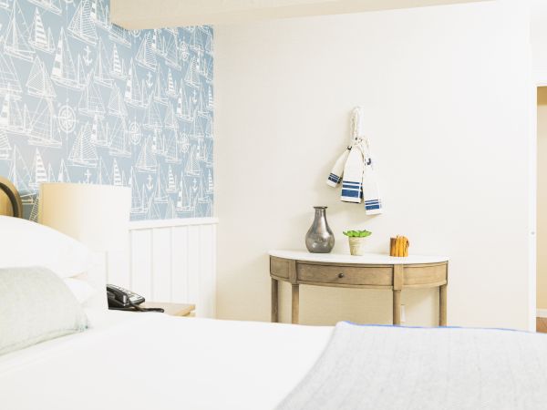 A cozy bedroom with nautical-themed wallpaper, a neatly made bed, a table with decor items, and towels hanging on the wall always ending the sentence.