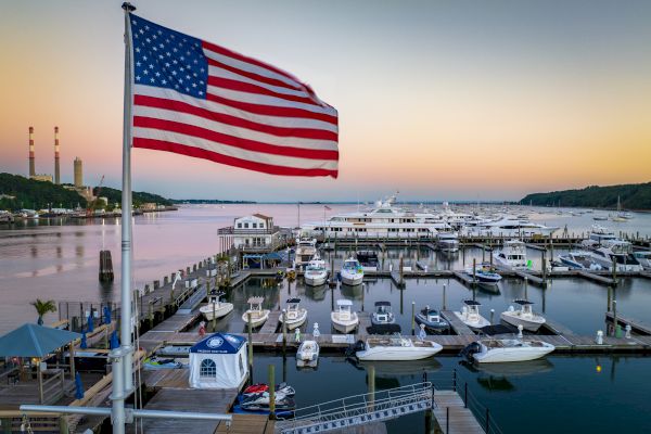 The image shows a marina with multiple boats docked, an American flag flying in the foreground, and a calm body of water at sunset.