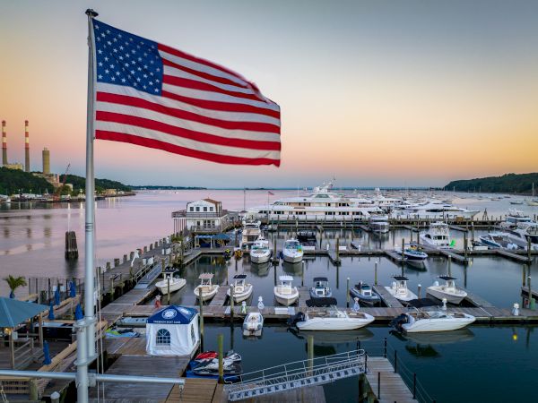 The image shows a marina with multiple boats docked, an American flag flying in the foreground, and a calm body of water at sunset.
