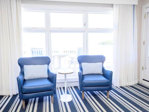 Two blue armchairs sit on a striped rug beside bright bay windows with curtains, creating a cozy sitting area.