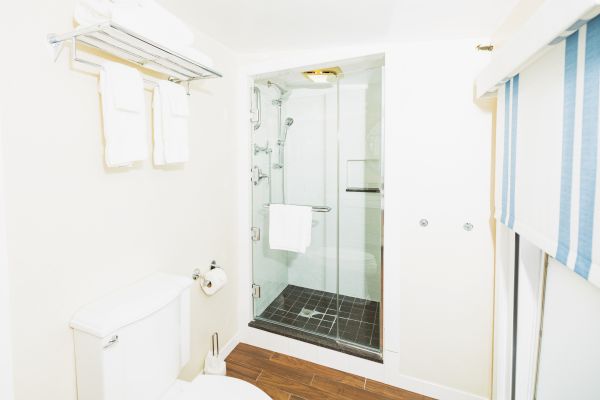 The image shows a clean bathroom with a glass-door shower, white towels on a rack, a toilet, and a window with blue and white striped curtains.