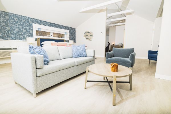 A modern living room with a gray sofa, armchair, wooden coffee table, and blue accent wall. There is also a bed in the background near the blue wall.