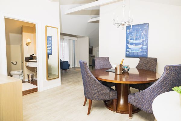 A dining area with a wooden table and blue upholstered chairs. There's a bathroom to the left and nautical-themed decor on the wall.