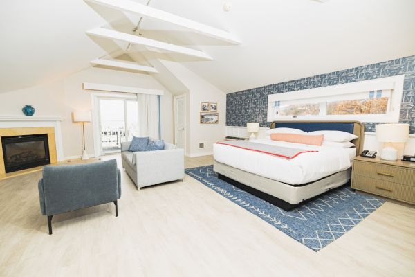 This image shows a modern bedroom with a large bed, blue accent wall, armchair, sofa, fireplace, and large windows for natural light.