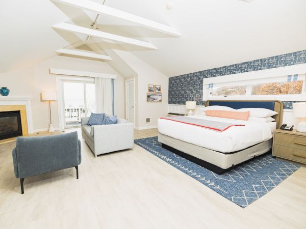 This image shows a modern bedroom with a large bed, blue accent wall, armchair, sofa, fireplace, and large windows for natural light.