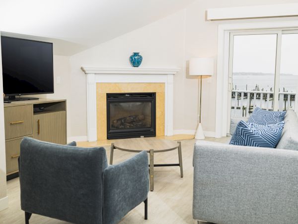 A modern living room features a fireplace, two armchairs, a sofa, a TV, a coffee table, and large windows with a view of a pier outside ending the sentence.