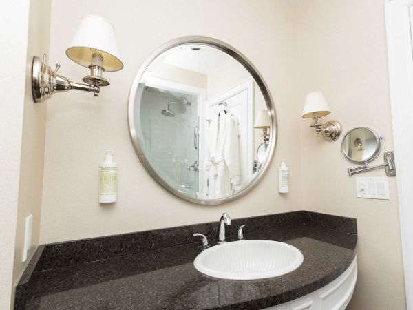 A modern bathroom vanity with oval mirrors, chrome wall lamps, toiletries, and a dark countertop sink.