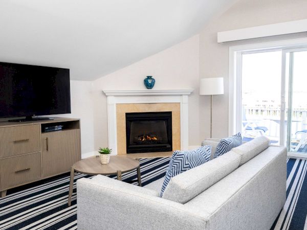 Bright living room with TV on a cabinet, a lit fireplace, a gray sofa with blue cushions, a striped rug, and a large window letting in light.