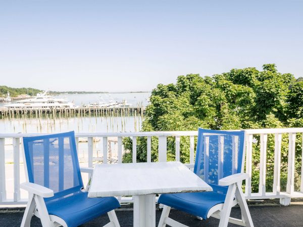Blue-and-white chairs on a balcony overlook a calm river, docks, and sailboats with green trees along the shore.