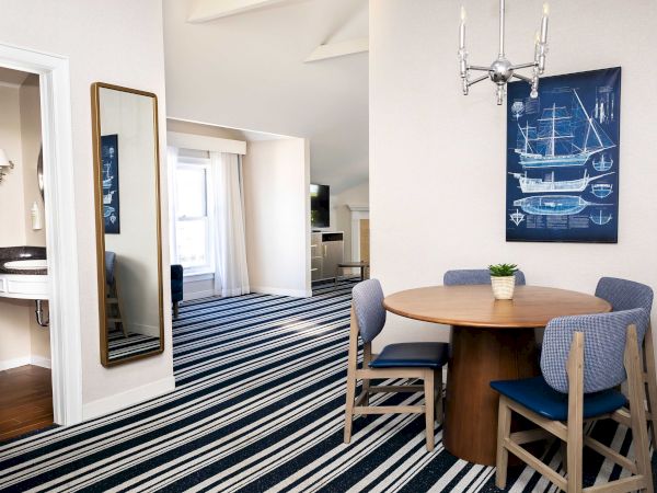 Dining nook with round wooden table, blue chairs, tall gold mirror, ship-illustration wall art, and chrome chandelier.