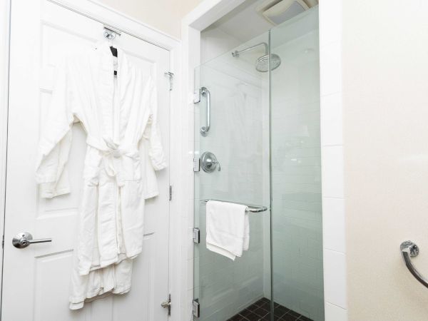 A hotel bathroom with a white robe hanging on the door, plus a glass shower and folded towels by the showerhead.
