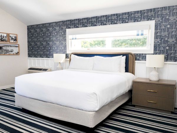 A bright hotel bedroom with a large white bed, two bedside lamps, patterned navy sailboat wallpaper, and a wide window with blinds.