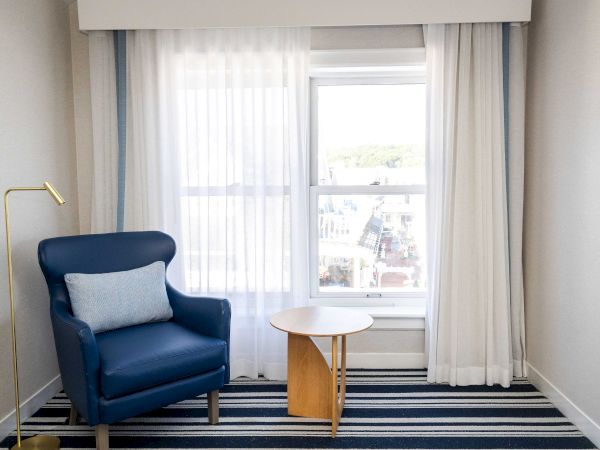 A bright room with a blue armchair, round wooden side table, and sheer curtains framing a large window.