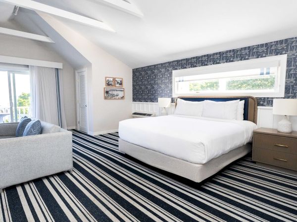 A bright hotel bedroom with a large bed, lamps, sofa by the window, striped carpet, and sailboat-pattern wallpaper.