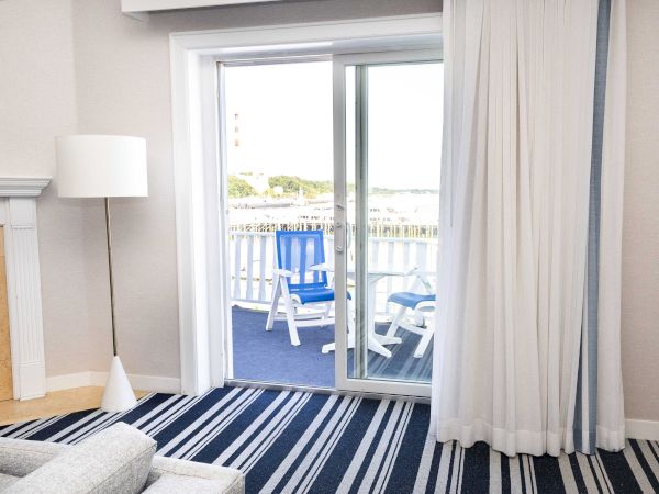 A bright hotel room opens onto a balcony with blue-and-white chairs overlooking the water and greenery outside.