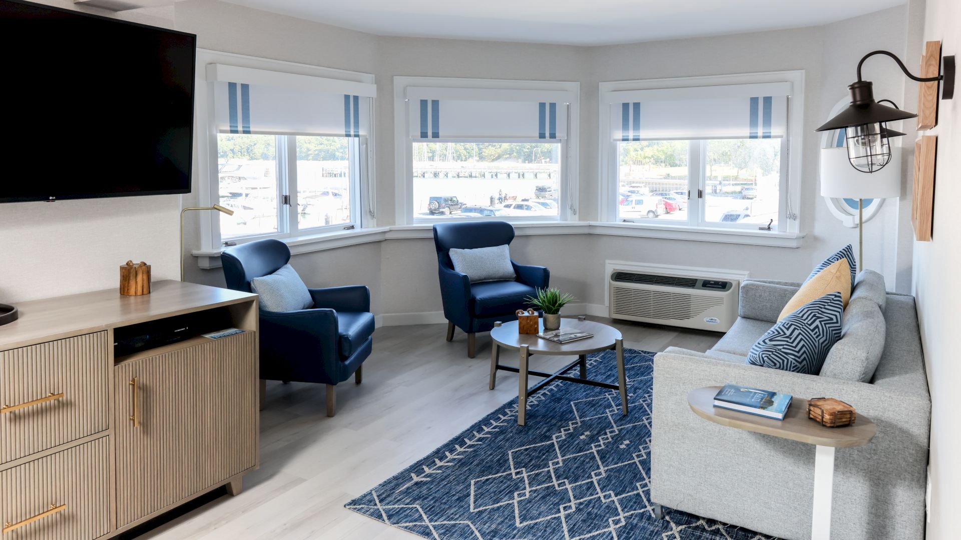 This image shows a modern living room with a sofa, blue armchairs, a coffee table, a wall-mounted TV, and large windows with striped blinds.