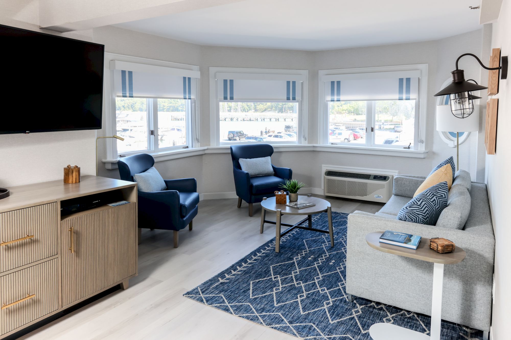 This image shows a modern living room with a sofa, blue armchairs, a coffee table, a wall-mounted TV, and large windows with striped blinds.