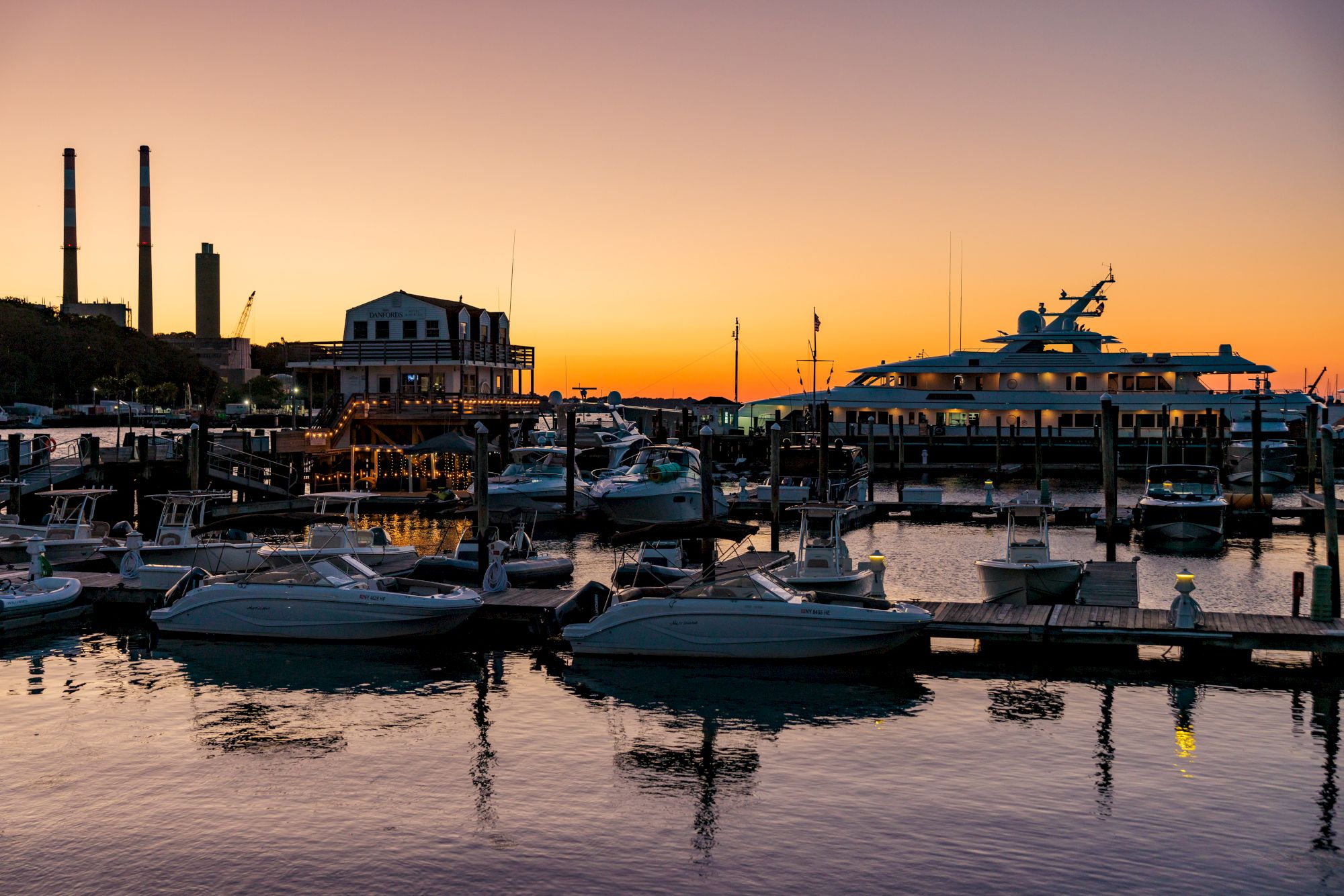 A serene marina at sunset with boats docked and buildings in the background, reflecting vibrant colors on the water's surface.