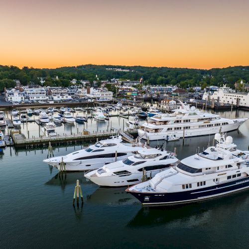 This image shows a marina with several yachts and boats docked, set against a backdrop of a small town and trees at sunset.