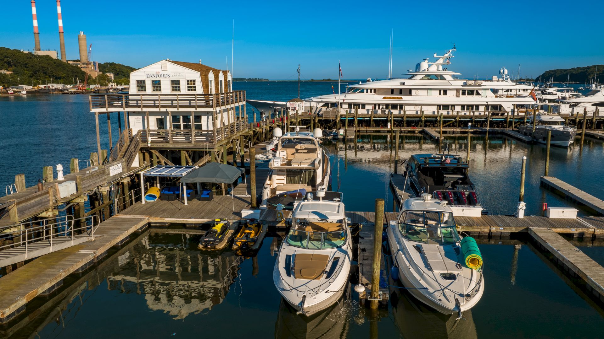 The image shows a marina with multiple boats and a large yacht docked, surrounded by wooden piers and a building, set against a clear blue sky.