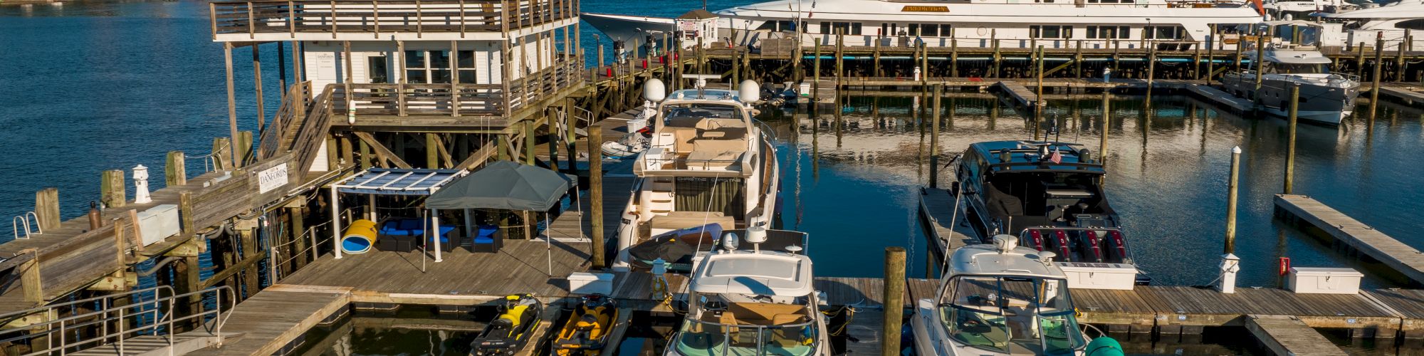 The image shows a marina with multiple boats and a large yacht docked, surrounded by wooden piers and a building, set against a clear blue sky.