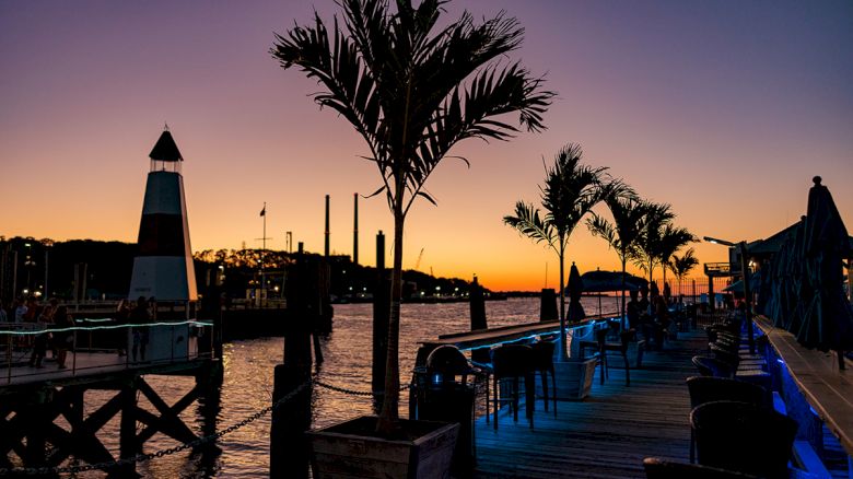 A sunset over a waterfront area with a lighthouse, palm trees, and an outdoor seating area along the dock, bathed in warm hues.