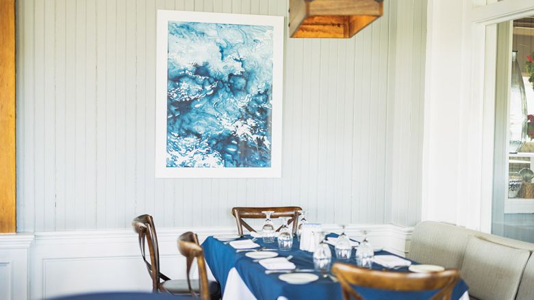 A dining area with a blue and white themed table setup, wooden chairs, a wall painting with blue tones, and a window showing an outside view.