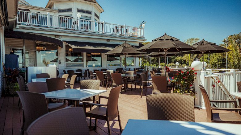 Outdoor dining area with tables, chairs, and black umbrellas on a deck. A two-story building with a balcony is in the background.