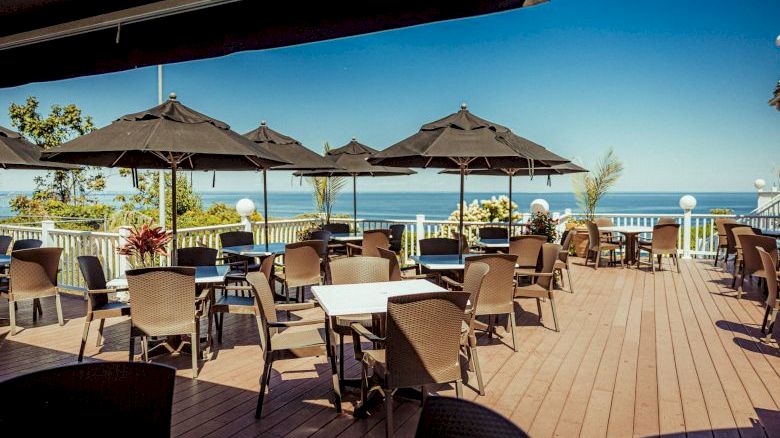 An outdoor dining area with tables and chairs, shaded by black umbrellas, overlooks a scenic ocean view under a clear blue sky.