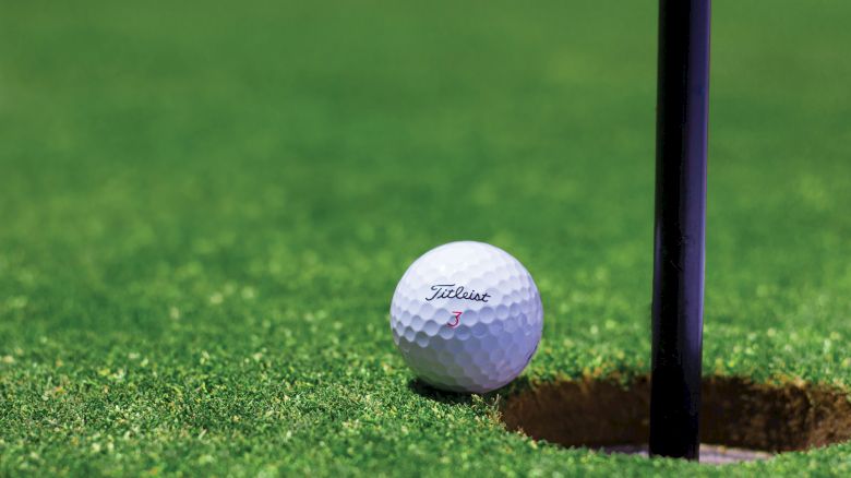 A close-up image of a golf ball resting near the edge of a hole on a green golf course, next to the flagstick.