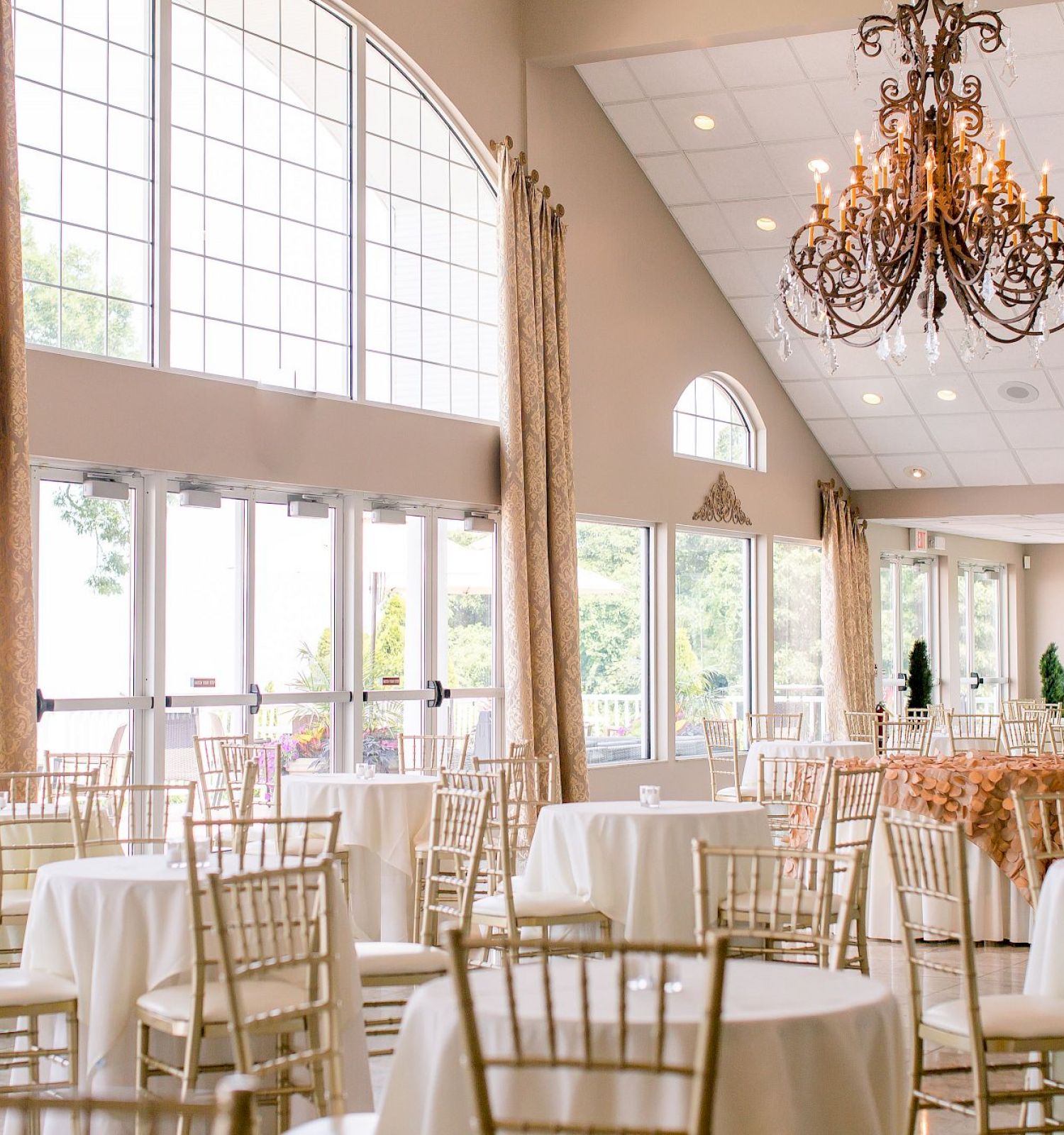 The image shows a spacious, elegantly decorated banquet hall with round tables, gold chairs, large windows, and a chandelier hanging from the ceiling.