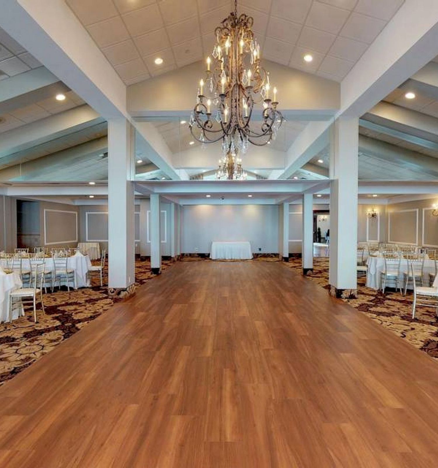 A spacious banquet hall with a chandelier, wooden flooring, and tables set with white tablecloths and chairs on either side of the room.