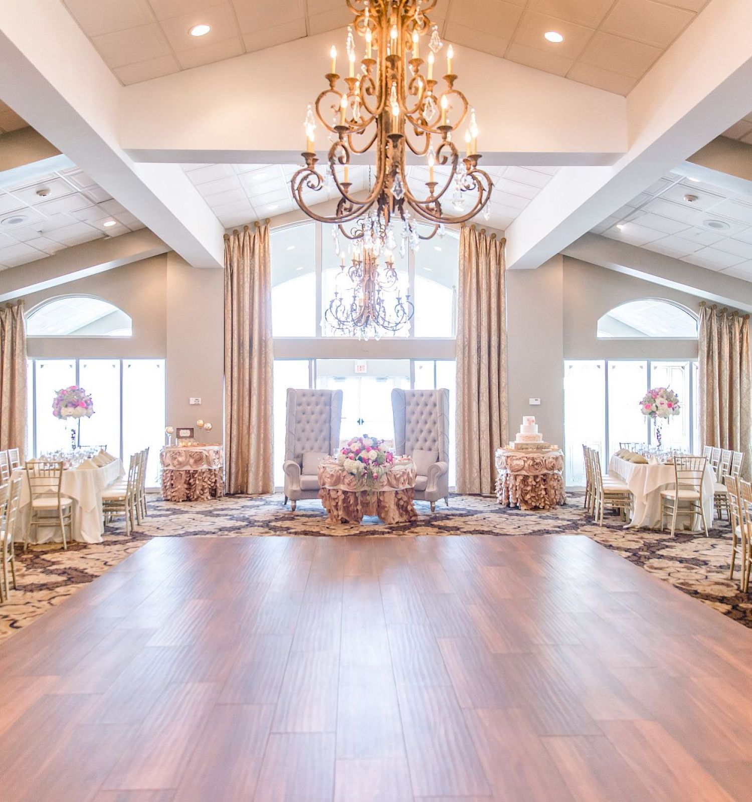 Elegantly decorated banquet hall with chandeliers, round tables with white tablecloths, floral centerpieces, and gold chairs, featuring large windows.