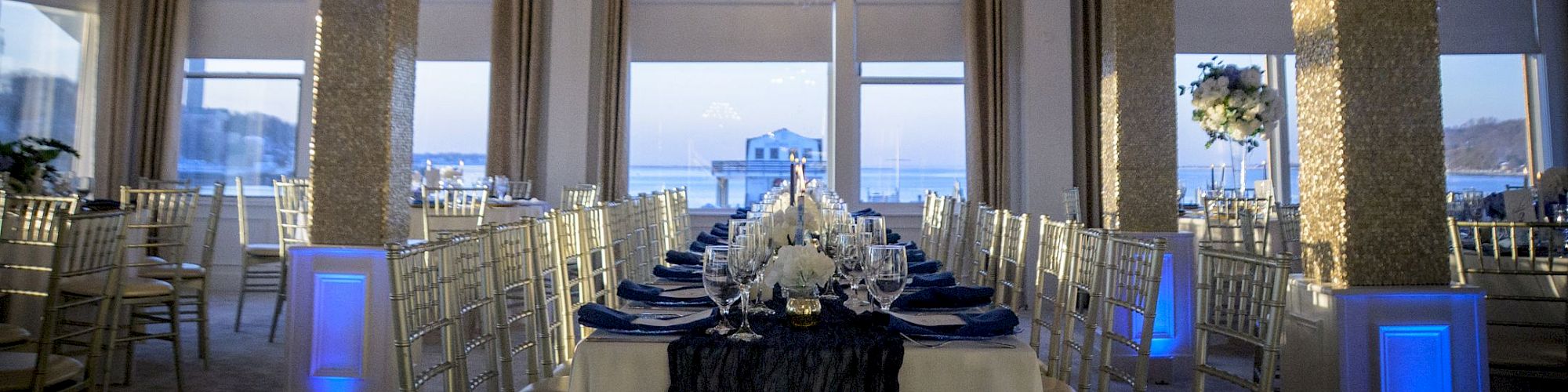 An elegant indoor banquet setup with long tables, white tableware, twinkling chandeliers, and blue lighting accents for a formal event.