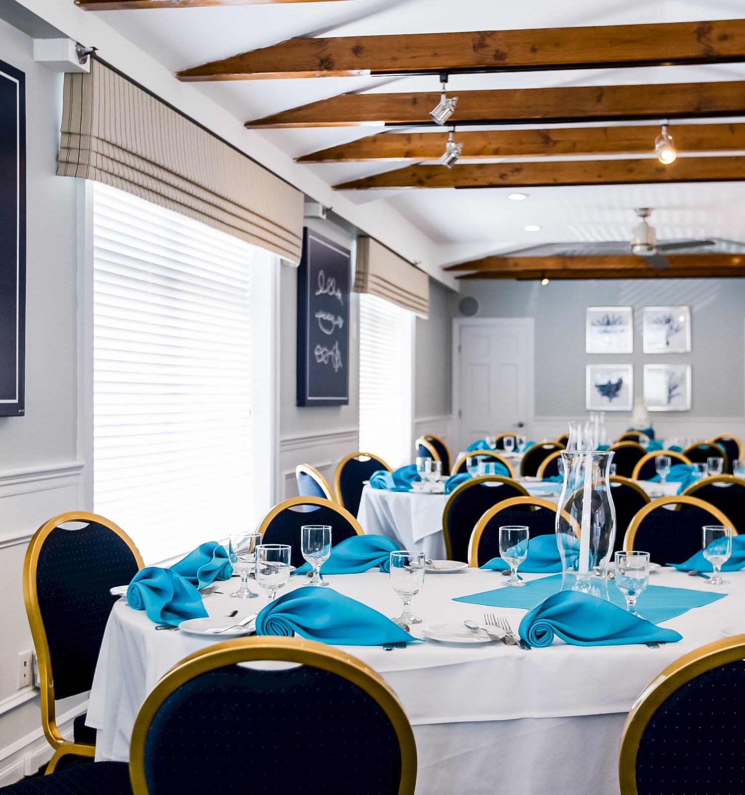 A bright dining room set for a formal event: white tablecloths, turquoise napkins, blue chairs, and chalkboard-style art on the walls.