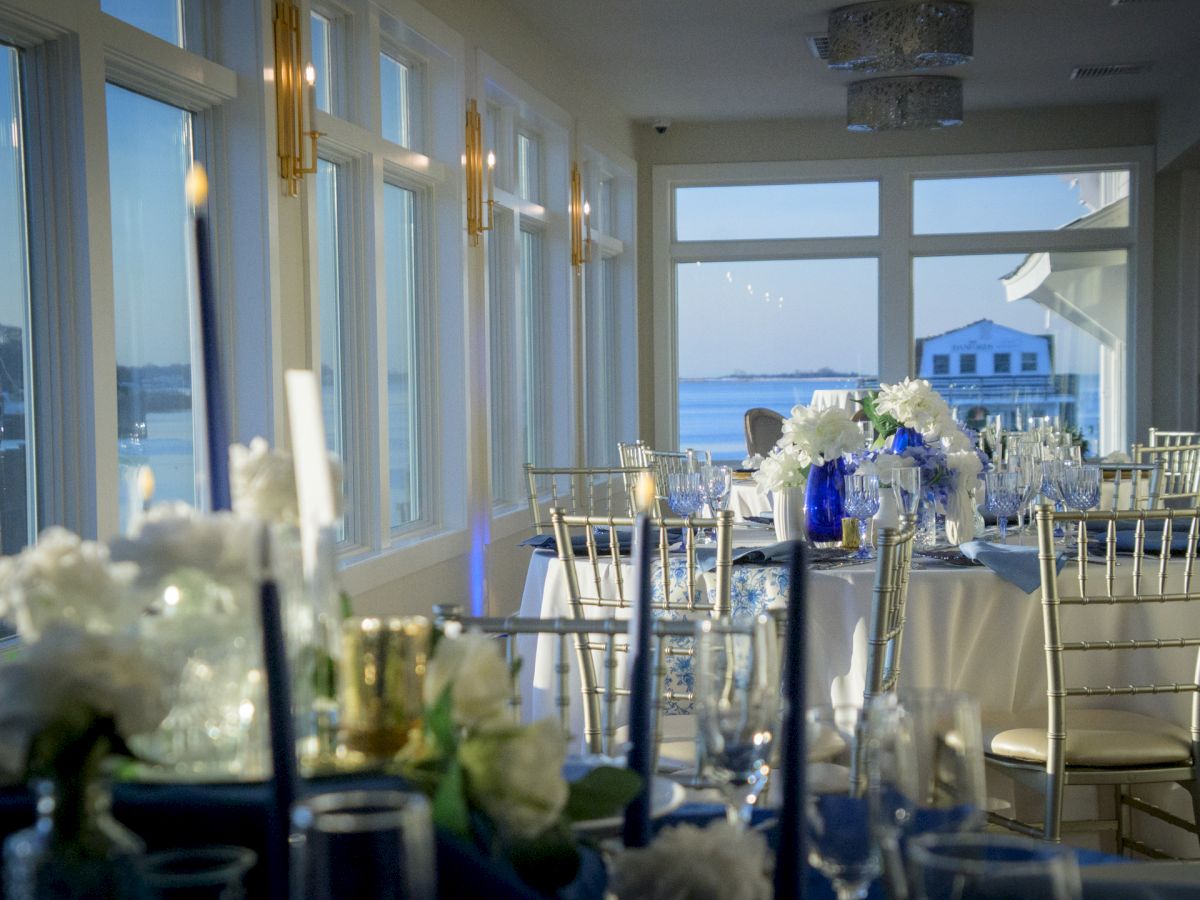 A bright seaside event space with round tables, white linens, silver chairs, floral centerpieces, candles, and large windows looking out to the water.