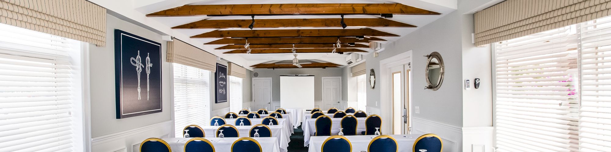 A classroom or meeting room setup with rows of blue and gold chairs, white-covered tables, a projector screen, ceiling fans, and artwork on the walls.