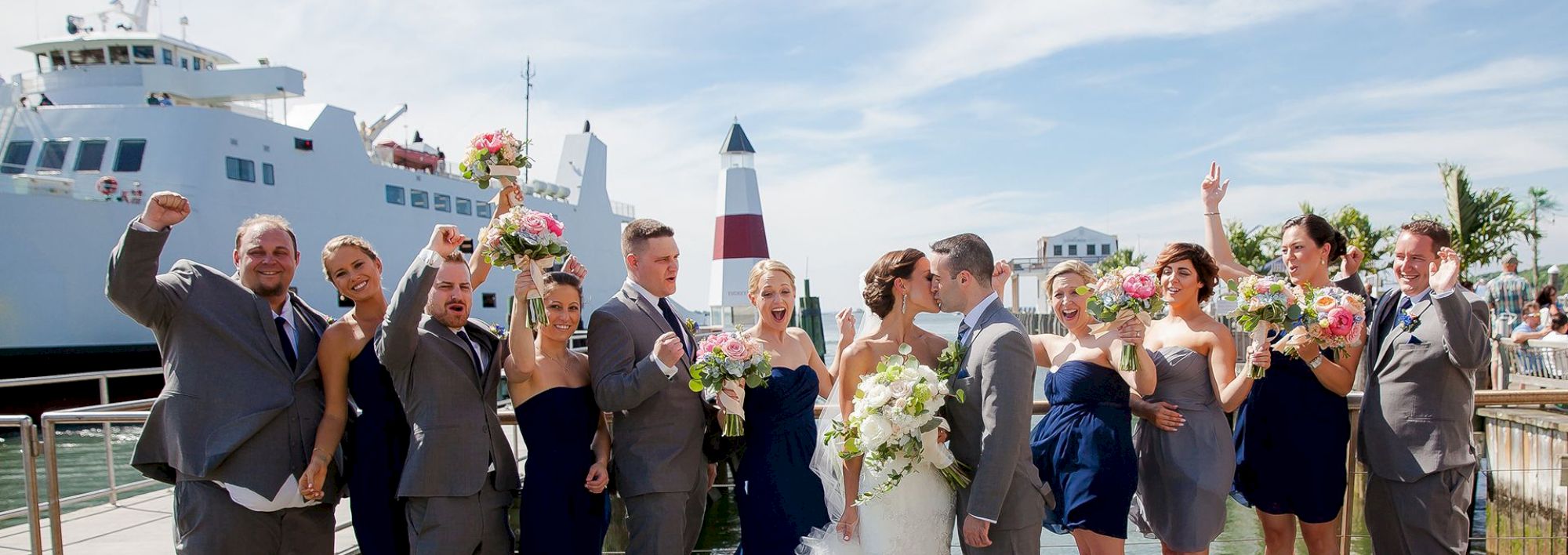 A wedding party celebrates near a large ferry boat; the bride and groom share a kiss while others cheer with bouquets raised.