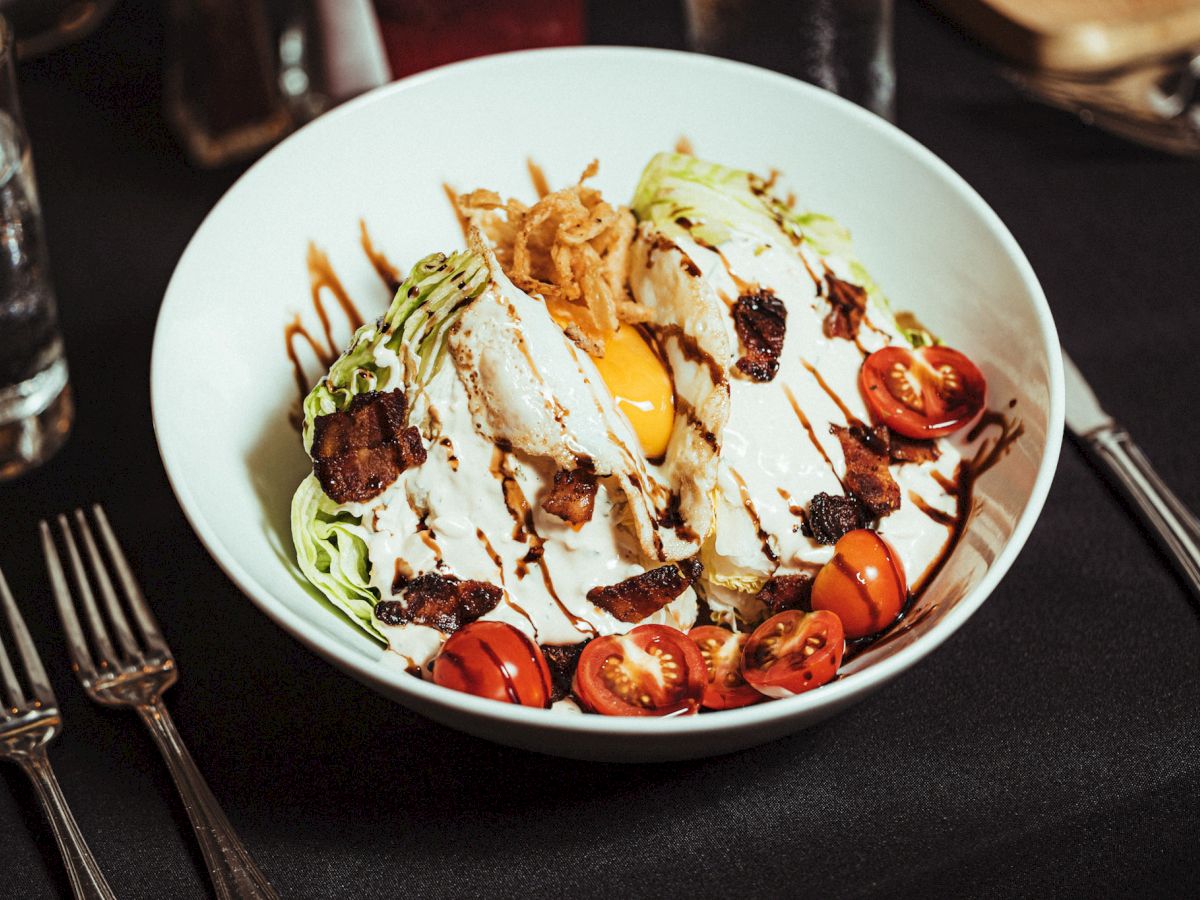 A plated dish with lettuce wedges, cherry tomatoes, bacon, a poached egg, and drizzled dressing sits on a black tablecloth with utensils.